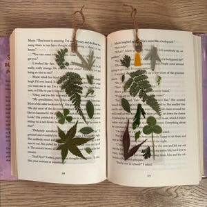 May include: Two book marks with pressed flowers and leaves. The bookmarks are made of brown string and have a yellow and green flower on the left bookmark and a green leaf on the right bookmark. The bookmarks are placed on an open book with text about a house and a bodyguard.