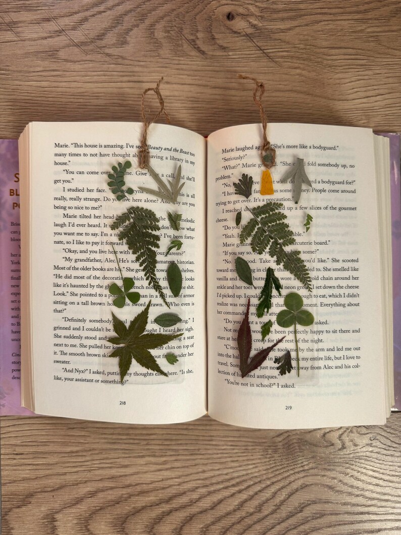 May include: Two book marks with pressed flowers and leaves. The bookmarks are made of brown string and have a yellow and green flower on the left bookmark and a green leaf on the right bookmark. The bookmarks are placed on an open book with text about a house and a bodyguard.