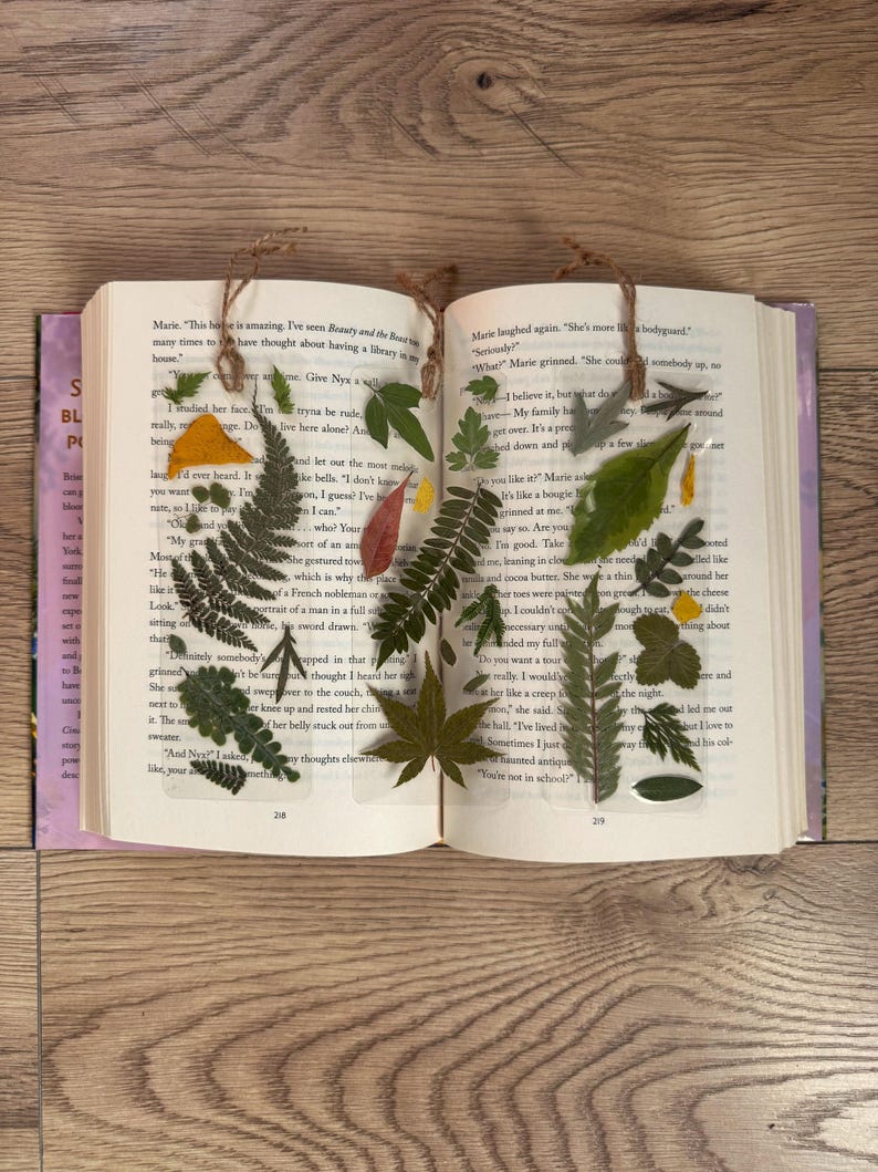 May include: A book with pressed leaves and flowers between the pages. The leaves are green, yellow, and red. The flowers are white and yellow. The book is open to a page with text about a house and a garden.