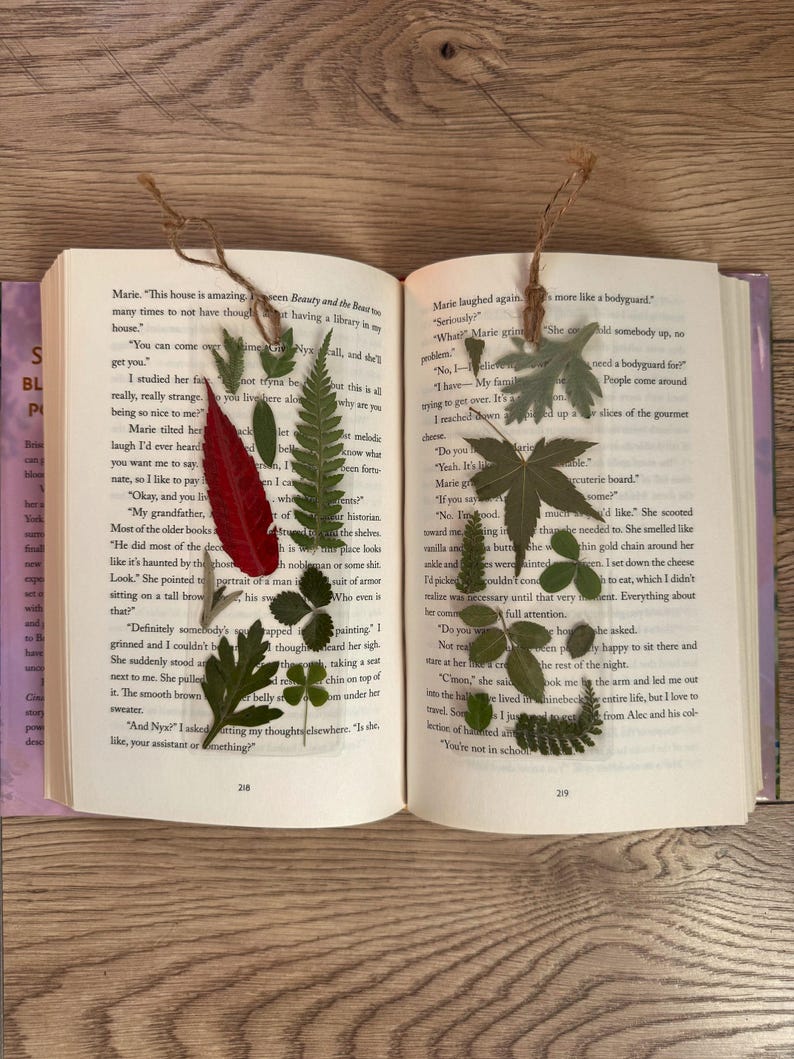 May include: An open book with pressed leaves and flowers as bookmarks. The book is open to pages with text about a house and a bodyguard. The leaves are green, red, and brown.