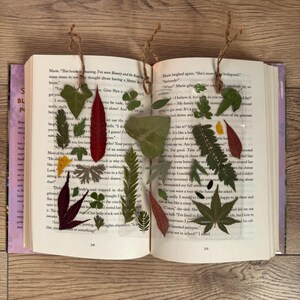 May include: Two clear resin bookmarks with pressed leaves and flowers. The bookmarks are on a book with text about a house and a bodyguard. The text on the book reads "This house is amazing. I've seen Beauty and the Beast many times to not think about having a library in the house." and "Marie laughed again. "She's more like a bodyguard."