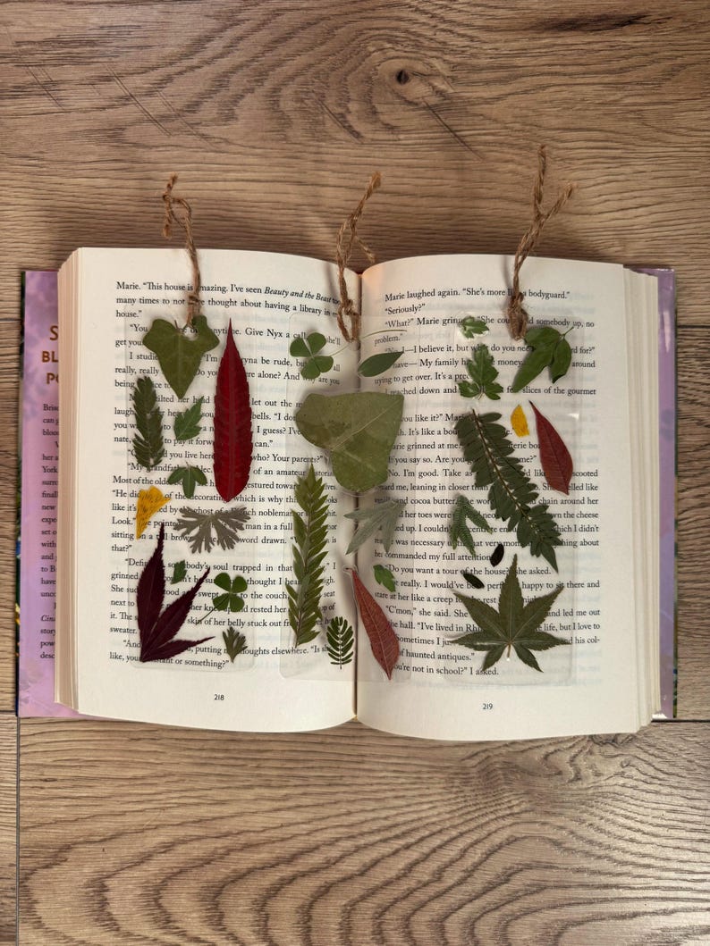 May include: Two clear resin bookmarks with pressed leaves and flowers. The bookmarks are on a book with text about a house and a bodyguard. The text on the book reads "This house is amazing. I've seen Beauty and the Beast many times to not think about having a library in the house." and "Marie laughed again. "She's more like a bodyguard."