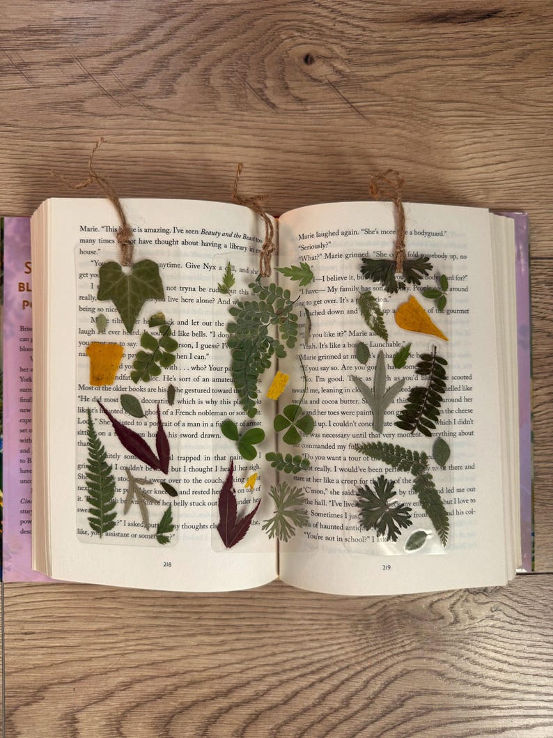 May include: Two handmade bookmarks with pressed flowers and leaves encased in clear resin. The bookmarks are attached to a book with twine. The bookmarks feature a variety of green leaves, yellow flowers, and a red leaf.