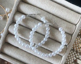 May include: A pair of large hoop earrings made of white faux pearls. The earrings are displayed in a jewelry box with other pearl jewelry.