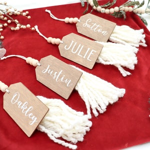 May include: Four wooden Christmas decorations with white tassels and names written in white on a red velvet background. The names are Oakley, Justin, Julie, and Sutton.