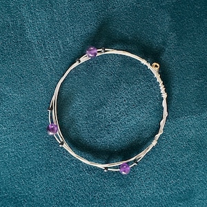 May include: A silver wire bracelet with three purple beads and small black beads. The bracelet is circular and appears to be handmade. The background is a dark teal color.