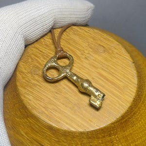 May include: A gold-colored key pendant with a leather cord rests on a wooden bowl. The key has a detailed design with two circular openings and a textured handle. The bowl is made of light brown wood.