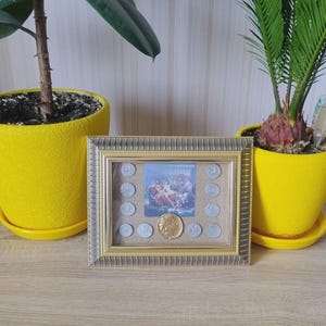 Puede incluir: Una imagen enmarcada en oro con una imagen vintage de una carrera de coches. La imagen está colocada sobre una mesa de madera junto a dos plantas en macetas amarillas.