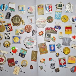 May include: A collection of vintage pins and badges, featuring various shapes and designs. The pins display a range of colors, including red, blue, gold, and white. Some pins have text, while others feature illustrations of flags, landmarks, and symbols.