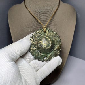 Puede incluir: Un colgante redondo de metal verde con intrincados diseños florales y un motivo circular central. El colgante está colgado de un cordón marrón.