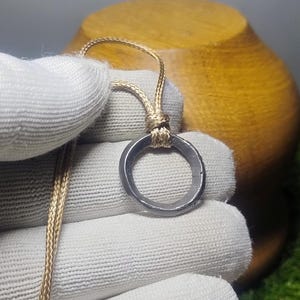 May include: A silver-colored pendant with a circular design, suspended from a beige cord. The pendant is held by a gloved hand, with a wooden object in the background. The cord is knotted at the top of the pendant.