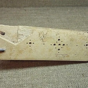 May include: A rectangular, off-white object with a series of small, drilled holes and a textured surface. The object has a geometric design on one end and is being touched by a metal tool.