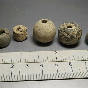 May include: Five antique clay beads, some with a hole in the centre, on a ruler with inch markings. The beads are various shades of brown and grey.