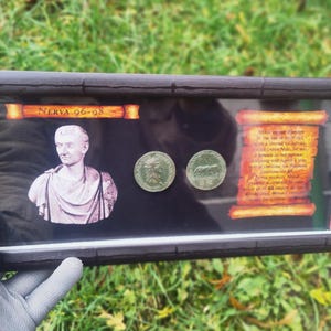 Pode incluir: Exibição emoldurada com um busto de Nerva, duas moedas antigas e um pergaminho com texto. A moldura é marrom escuro e o fundo é preto. O texto no pergaminho é laranja desbotado. As moedas são verdes.
