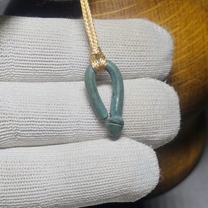 Puede incluir: Primer plano de un pequeño colgante turquesa con diseño de lazo y base redondeada. El colgante está sujeto a un fino cordón dorado. El fondo es una superficie blanca texturizada.