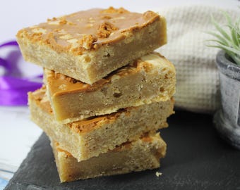 May include: A stack of four homemade caramel squares with a light brown color and a slightly crispy texture. The squares are arranged on a black slate surface.