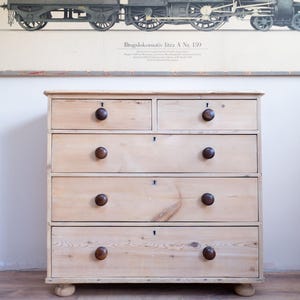 May include: A pale wooden chest of drawers with five drawers and dark brown, round knobs. The chest is positioned against a wall featuring a vintage train illustration. The drawers have keyholes, and the chest rests on small, rounded feet.