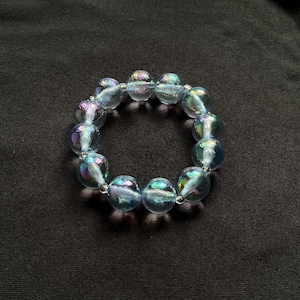 May include: A bracelet made of iridescent, clear glass beads. The beads are round and strung together on a clear elastic cord. The beads have a blue and purple sheen, and the bracelet is set against a black background.