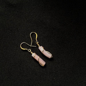 May include: A pair of dangle earrings with pale pink, cylindrical stones wrapped in gold-coloured wire. The earrings have gold-coloured ear wires against a black background. The earrings are a type of jewellery.
