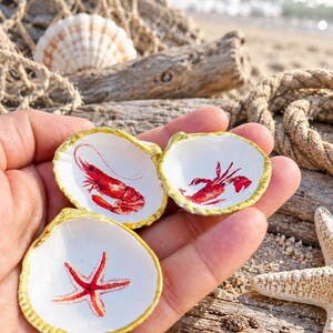 May include: Three small, white, concave seashells with gold-painted edges. Each shell features a detailed, colorful illustration on its interior: one depicts a red shrimp, one a red lobster, and one a red starfish, all centered on the white surface.