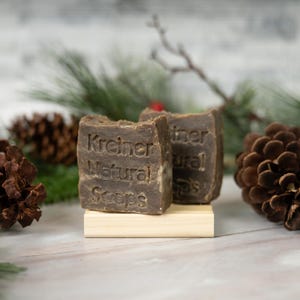 May include: Two brown bars of soap with the text "Kreiner Natural Soaps" are displayed on a small wooden stand. The background includes pine cones, evergreen branches, and a blurred white surface.