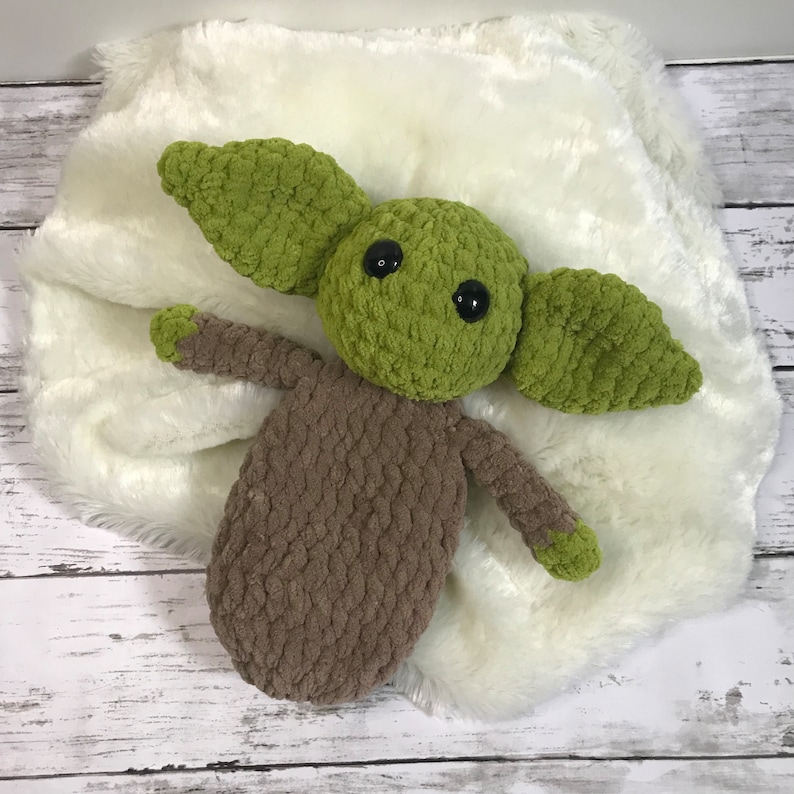 May include: A green and brown crocheted plush toy resembling a popular fictional character. The toy is lying on a white fuzzy blanket.