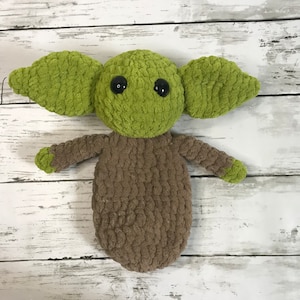 May include: A green and brown crocheted plush toy resembling a character from the Star Wars franchise. The toy has large ears, big eyes, and a small body.
