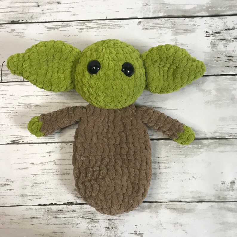 May include: A green and brown crocheted plush toy resembling a character from the Star Wars franchise. The toy has large ears, big eyes, and a small body.