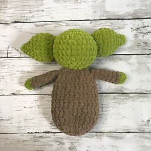 May include: A green and brown crocheted plush toy resembling a character from the Star Wars franchise. The toy is lying on a white wooden surface.