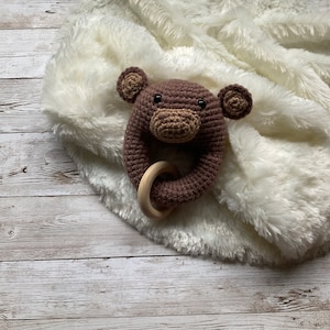 May include: A brown crocheted monkey rattle with a wooden ring. The rattle is on a white fluffy blanket.