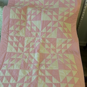 May include: A rectangular quilt featuring a geometric pattern of alternating light pink and off-white triangular fabric patches. The quilt is finished with a solid light pink border and displays visible linear stitching throughout the surface.