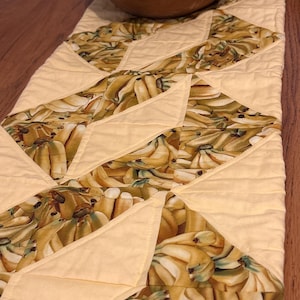 May include: A yellow table runner with a geometric pattern of yellow and brown banana bunches. The runner is made of fabric and has a quilted texture.