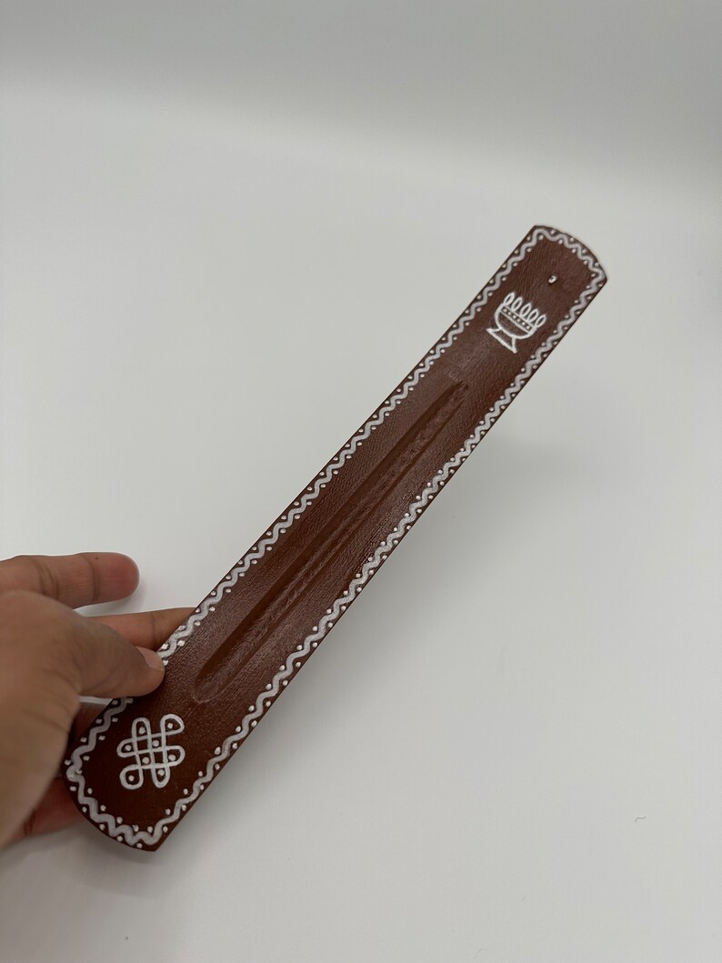 Op de afbeelding: Bruine houten wierookbrander met witte geverfde ontwerpen. De ontwerpen omvatten een gestileerde bloem en een knoop.