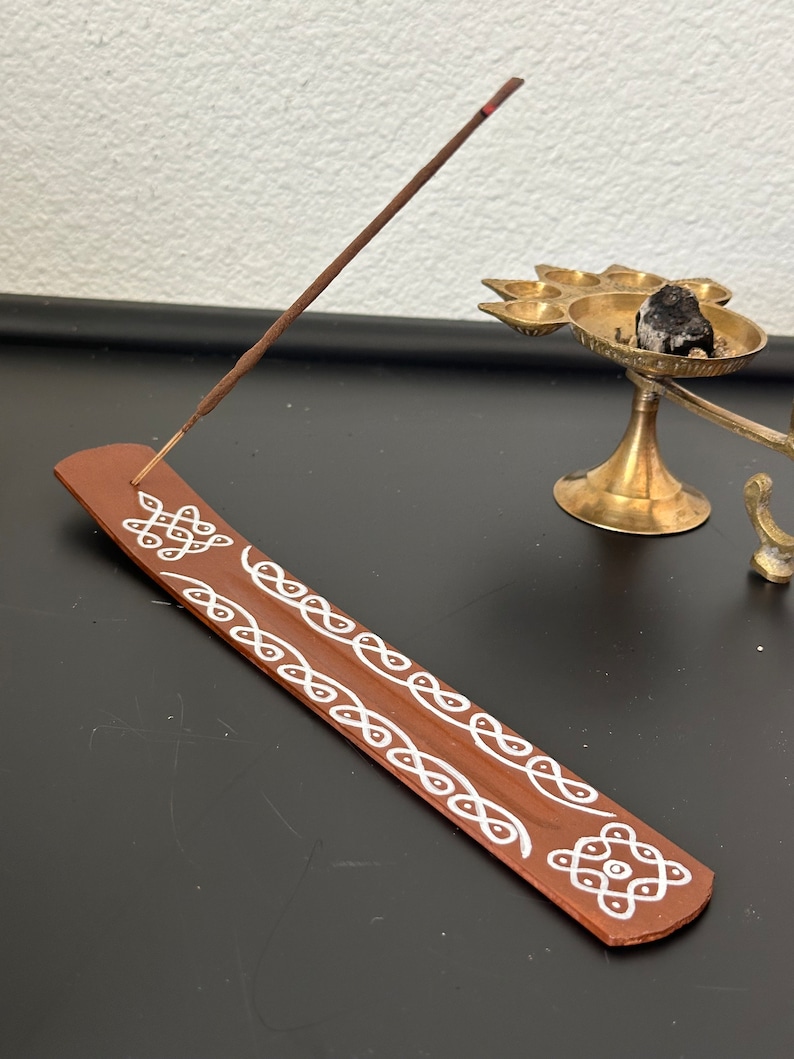 Op de afbeelding: Een bruine houten wierookstokhouder met wit geschilderde ontwerpen. De houder is versierd met een herhalend patroon van cirkels en lussen. Een wierookstokje brandt in de houder.