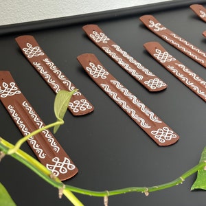 Op de afbeelding: Een set van acht bruine wierookstokjes met witte geometrische ontwerpen. De stokjes zijn in een rij op een zwart oppervlak gerangschikt.