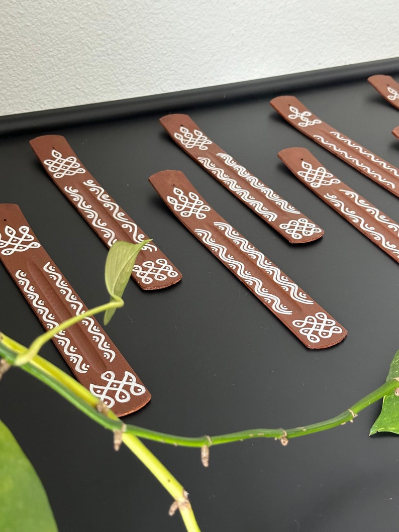Op de afbeelding: Een set van acht bruine wierookstokjes met witte geometrische ontwerpen. De stokjes zijn in een rij op een zwart oppervlak gerangschikt.