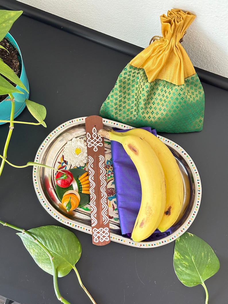 Op de afbeelding: Een zilveren dienblad met een paarse doek, twee gele bananen, een witte bloem, een rode en groene chilipeper, een klein oranje en groen voorwerp en een bruine houten wierookstok met witte markeringen. Het dienblad is versierd met een kleurrijke rand.