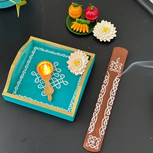 Op de afbeelding: Een tealblauwe houten dienblad met witte geschilderde ontwerpen en een messing olielamp. Het dienblad is versierd met een witte bloem en een klein bord met groen, geel en rood fruit. Een bruine houten wierookstok met witte geschilderde ontwerpen is ook te zien.