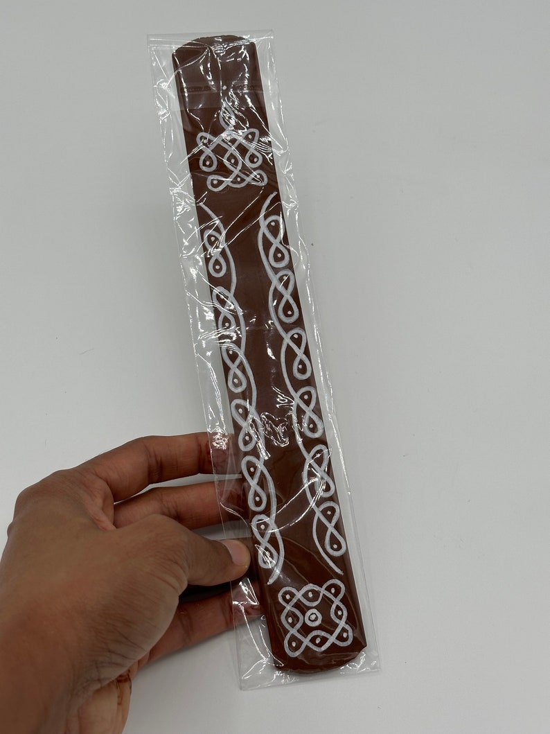 Op de afbeelding: Een bruine wierookstok met witte decoratieve ontwerpen. De stok is verpakt in doorzichtig plastic.
