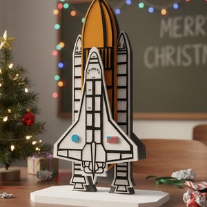 May include: A wooden space shuttle Christmas decoration with a white and black design, orange top, and American flag detail. A small Christmas tree and the words "MERRY CHRISTMAS!" are in the background.