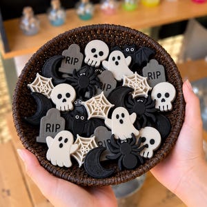 May include: A dark brown woven basket filled with small Halloween-themed decorations including white skulls, grey tombstones with the text RIP, white spiderwebs, black spiders, black crescent moons, and white ghosts.