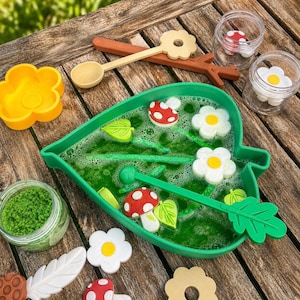 May include: A green leaf-shaped basin filled with green soapy water containing floating white flower and red mushroom shapes. Includes a green leaf-shaped stirring wand, a tan flower-shaped wand, a jar of green granules, and two clear jars with small mushroom and flower pieces.