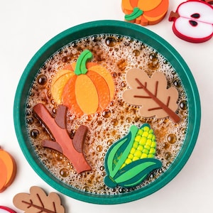 May include: A dark green circular basin filled with brown liquid and bubbles, containing floating autumn-themed shapes including an orange pumpkin, a brown oak leaf, a brown twig, and a yellow corn cob with green husks.