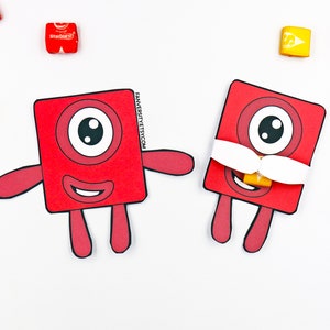 May include: Two red paper cutout monsters with one eye and a mouth. The monster on the right has a white paper mustache and a yellow candy bar for a nose.
