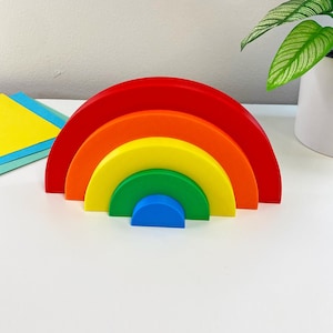 Può includere: Un giocattolo impilabile a forma di arcobaleno in legno con archi rossi, arancioni, gialli, verdi e blu. Il giocattolo è progettato per essere incastrato, creando una forma ad arcobaleno. Il giocattolo è su una superficie bianca.