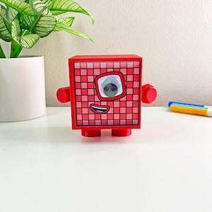May include: Red toy robot with a single eye and a smiling mouth. The robot has a grid pattern on its body and is standing on a white surface.