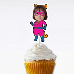 May include: A cupcake with white frosting and a pink, blue, and yellow superhero cupcake topper with a girl's face. The superhero is wearing a mask and has cat ears.