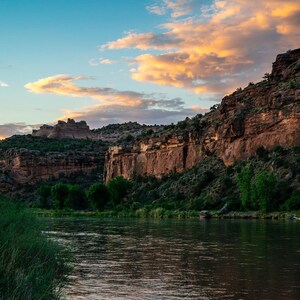 May include: A scenic view of a river flowing through a canyon with red rock cliffs and lush green vegetation. The sky is a vibrant blue with fluffy clouds reflecting in the water.
