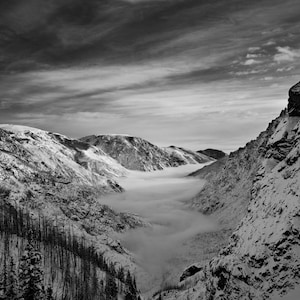 May include: A black and white photograph of a snow-covered mountain valley with a thick layer of fog in the valley. The fog is so thick that it obscures the bottom of the valley. The mountains are steep and rocky, and the sky is overcast.