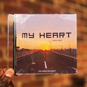 Può includere: Una custodia per CD con una foto di un tramonto di una strada che porta a un ponte. Il testo sulla custodia recita "My Heart Since 1957" e "You Make Me Happy".